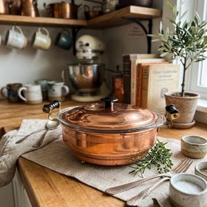 May include: A round copper casserole dish with a glass lid, featuring two brass handles with black accents and a black knob on the lid. The exterior of the pot has horizontal ribbed detailing. It sits on a linen cloth alongside silver forks.