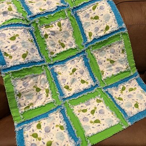 May include: A blue and green patchwork quilt with a white fabric featuring a pattern of sea turtles, jellyfish, and fish. The quilt is made with a ruffled edge.