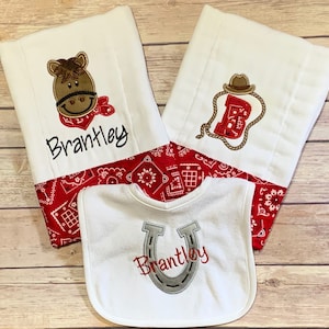 May include: A set of three white baby burp cloths with red bandana trim. One cloth features a brown horse with a red bandana, another features a red letter B with a brown cowboy hat and rope, and the third cloth is a bib with a gray horseshoe and the name "Brantley" embroidered on it.