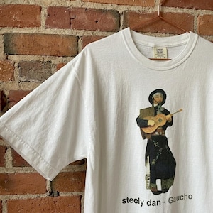 Puede incluir: Camiseta blanca con un gráfico de una persona tocando la guitarra, vestida con abrigo y sombrero negros. El texto "steely dan - Gaucho" está impreso en la parte inferior. La camiseta está colgada en una percha color cobre.