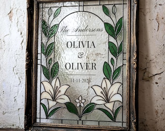 Può includere: Un pannello rettangolare in vetro colorato in una cornice ornata color bronzo scuro con fiori di giglio bianchi e foglie verdi. Il vetro mostra il testo The Andersons, OLIVIA & OLIVER e la data 11-11-2026 in lettere nere su superficie ruvida.