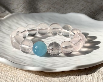 Op de afbeelding: Een kralenarmband met doorschijnende, lichtroze bolvormige kralen en één enkele, ondoorzichtige, helder hemelsblauwe bolvormige kraal. De kralen zijn in een cirkel gerangschikt en hebben een glad, gepolijst oppervlak dat het licht weerkaatst.