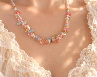 Op de afbeelding: Een ketting met een streng kleine, onregelmatige, gepolijste edelsteensplinters in roze, gele en lichtblauwe tinten, geaccentueerd met kleine goudkleurige kraaltjes en geregen aan een fijne, lichtgekleurde kralenketting.