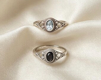 Op de afbeelding: Twee zilveren ringen met een ovale zetting met kralenranden en opengewerkte metalen details. De bovenste ring bevat een lichtblauwe gefacetteerde edelsteen, terwijl de onderste ring een gladde, donkerzwarte ovale steen in het midden heeft.