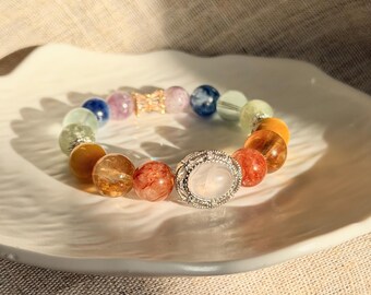 Op de afbeelding: Een kralenarmband met een verscheidenheid aan gepolijste, veelkleurige edelsteenbollen in tinten blauw, paars, groen, geel en oranje. De armband bevat een centrale zilverkleurige ovale bedel met een witte steen en een klein vlinderaccent.