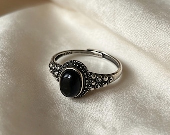 Op de afbeelding: Een verstelbare zilveren ring met een centrale ovale zwarte steen in een kraalrand. De band heeft een textuur met een stippenpatroon aan de zijkanten en is aan de binnenzijde voorzien van de tekst S925 in het metaal gegraveerd.