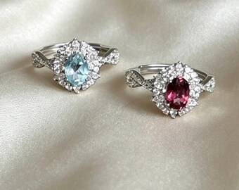 Op de afbeelding: Twee zilverkleurige ringen met ovaal geslepen middenstenen, één lichtblauw en één donkerroze. Elke ring is omringd door een halo van kleine heldere steentjes en heeft een gedraaide band met extra kleine heldere steentjes aan de zijkanten.
