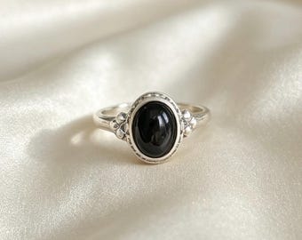 Op de afbeelding: Een zilveren ring met een centrale ovaalvormige zwarte steen. De zetting is voorzien van een kralenrand rondom de steen en kleine decoratieve metalen bolletjes aan weerszijden van de ringband, die overgaat in een gladde, gepolijste zilveren schacht.