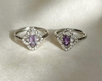 Op de afbeelding: Twee zilverkleurige ringen met een vierbladig bloemmotief bezet met kleine heldere steentjes. De linkerring heeft een rechthoekige lichtpaarse middensteen, terwijl de rechterring een ovale dieppaarse middensteen bevat.