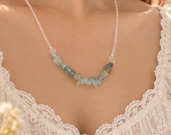 Op de afbeelding: Een ketting met een streng kleine, doorzichtige kralen die overgaat in een middengedeelte van onregelmatige, getrommelde edelsteensplinters in lichtblauwe, groene en paarse tinten, gedragen op een kledingstuk met witte kanten afwerking.