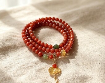 Op de afbeelding: Een armband met meerdere strengen bestaande uit kleine, ronde, rood-oranje kralen. De armband is voorzien van twee kleine groene kralen, één gele kraal en een goudkleurige kettinghanger met een gele druppelvormige kraal en twee kleine rode kralen.