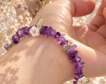 Op de afbeelding: Een armband met onregelmatige paarse amethist chip kralen, afgewisseld met kleine witte bloemvormige bedels en doorschijnende groene bladvormige accenten. De kralen zijn aan elkaar geregen tot een doorlopende band van violette stenen.