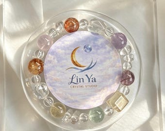 Op de afbeelding: Een cirkelvormige kralenarmband met een verscheidenheid aan doorschijnende en halfedelstenen in tinten van helder, amber, paars en groen. Een ronde kaart in het midden toont een maansikkel-logo en de tekst Lin Ya CRYSTAL STUDIO.