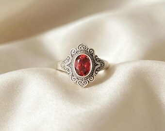 Op de afbeelding: Een zilveren ring met een ovaal geslepen dieprode edelsteen, gezet in een decoratief frame met ingewikkelde krulmotieven en kraalafwerking. De metalen band heeft een gepolijste afwerking en de zetting is gevormd met symmetrische curven.