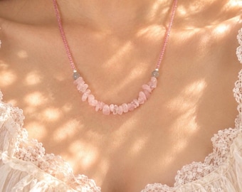 Op de afbeelding: Een ketting met een streng kleine roze kralen verbonden aan een middengedeelte van onregelmatige, getrommelde lichtroze steensplinters. Twee kleine, ronde, doorschijnende grijze kralen bevinden zich tussen de roze steentjes en de kralen.