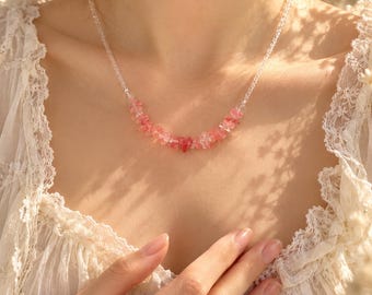 Op de afbeelding: Een ketting bestaande uit een streng heldere, transparante kralen die overgaat in een cluster van kleine, onregelmatige, roze en rode edelsteensplinters die centraal aan de voorzijde bij de hals rusten.