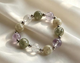 Op de afbeelding: Een kralenarmband met afwisselend ronde gepolijste stenen in saliegroen, doorschijnend helder en licht lavendel, gescheiden door kleine metallic zilverkleurige tussenkralen die in een cirkelvormig patroon zijn gerangschikt.
