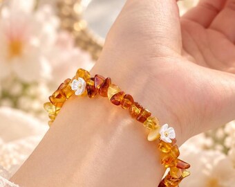 Op de afbeelding: Een armband bestaande uit onregelmatige, gepolijste barnstenen in tinten honing, goud en donkerbruin, afgewisseld met twee kleine, witte, vijfbladige bloembedels met piepkleine gele hartjes, geregen aan een elastisch koord.