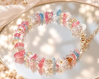 Op de afbeelding: Een kralenarmband met onregelmatig gepolijste schilfers van roze, blauwe en gele edelstenen, afgewisseld met kleine witte bloemvormige kralen. De armband is voorzien van een goudkleurige kettingverlenger en een karabijnsluiting.