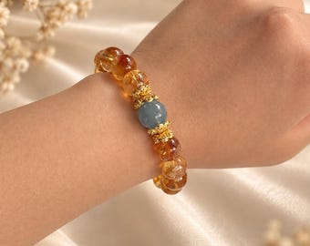 Op de afbeelding: Een kralenarmband met doorschijnende amberkleurige bolvormige kralen en een enkele centrale matte blauwe kraal. De kralen worden gescheiden door kleine, ingewikkelde goudkleurige metalen tussenstukjes met een bloemachtig ontwerp.