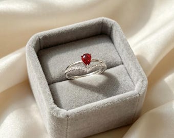 Op de afbeelding: Een zilverkleurige ring met een peervormige rode edelsteen in een zetting, vergezeld van een V-vormige band ingelegd met kleine heldere steentjes. De ring wordt gepresenteerd in een vierkant, met grijs fluweel bekleed sieradendoosje.