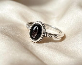 Op de afbeelding: Een zilveren ring met een ovaalvormige zwarte steen in een decoratieve zetting met een kralenrand. De ringband heeft een textuur met een stippenpatroon aan de zijkanten, wat overgaat in een gladde, gepolijste afwerking aan de achterzijde.