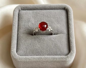 Op de afbeelding: Een zilverkleurige ring met een centrale, ronde en gepolijste dieprode edelsteen. De ringband is aan beide zijden van de steen versierd met kleine, heldere, fonkelende steentjes in een bladvormig metalen ontwerp.