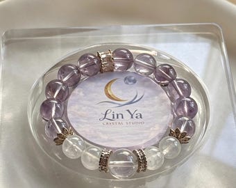 Op de afbeelding: Een kralenarmband met doorschijnende paarse en heldere bolvormige stenen. Het ontwerp bevat goudkleurige metalen tussenstukjes en decoratieve bloemvormige accenten. Een ronde kaart in het midden toont de tekst Lin Ya CRYSTAL STUDIO.