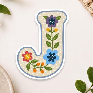May include: A die-cut sticker in the shape of the letter J, featuring a beige background with white polka dots and a blue border. The letter is decorated with a vertical floral pattern including a purple flower, a blue flower, and a red flower.