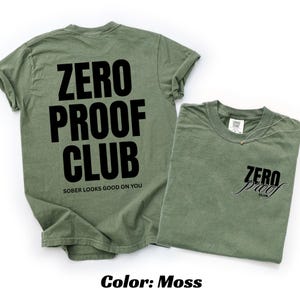May include: Two moss-colored t-shirts. One shirt displays "ZERO PROOF CLUB" in large black letters on the back, with "SOBER LOOKS GOOD ON YOU" below. The other shirt has "ZERO proof CLUB" in script on the front. The color is "Moss".