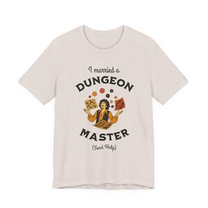 May include: A light heather gray t-shirt with the text "I married a DUNGEON MASTER (Send Help)". The graphic features a person juggling dice and game cards. The shirt is a short-sleeved crew neck.