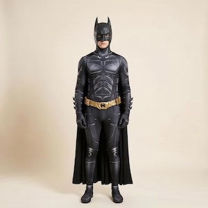 Puede incluir: Un disfraz de superhéroe negro de cuerpo completo que presenta una pieza pectoral moldeada, guanteletes integrados y una capucha a juego con orejas puntiagudas. El traje incluye un cinturón utilitario dorado con un emblema central y una capa negra larga.