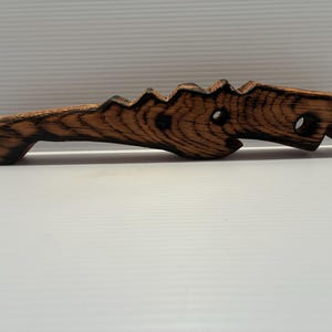 May include: A handcrafted wooden bottle opener with a unique, stylized design. The opener is made of dark brown wood with visible grain and a textured surface. It features a handle and a decorative, jagged edge.