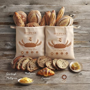 May include: Two beige beeswax bread bags with brown text and graphics, including a bee icon, wheat stalks, and the phrase Love Every Slice. The bags are filled with various loaves of bread, with sliced sourdough, multigrain bread, and a croissant arranged in front.