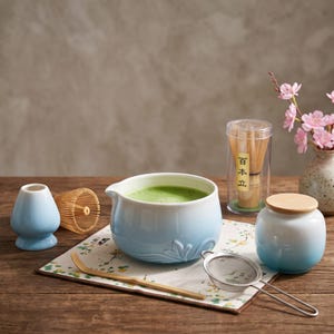 May include: A matcha tea set featuring a light blue gradient ceramic bowl filled with green tea, a matching lidded jar, a light blue whisk holder, a bamboo whisk in a clear case with Japanese text, a bamboo scoop, and a metal tea strainer.