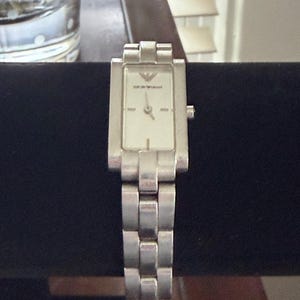 May include: Silver-toned rectangular wristwatch with a white face and silver band. The watch face features the brand name "Emporio Armani". The watch is displayed on a black surface.