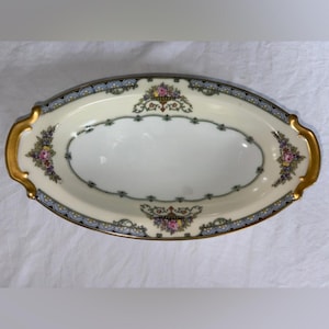 May include: An oval serving dish with a cream-colored interior and gold handles. The rim features a blue and gold design with floral accents in pink, yellow, and purple. The dish is approximately 12 inches long.