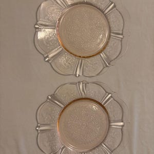 Puede incluir: Dos platos de cristal transparente con un centro rosa y un motivo floral decorativo. Los platos tienen un borde festoneado y están dispuestos uno al lado del otro. Los platos son probablemente para servir o exhibir.