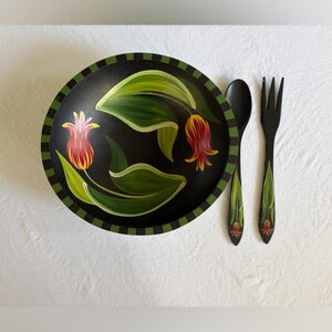 May include: A black wooden bowl, spoon, and fork set. The bowl features a floral design with red and yellow flowers and green leaves. The spoon and fork are black with matching floral accents. The set is on a white surface.