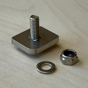 Könnte beinhalten: Ein Satz Metallbeschlagteile, darunter eine Schraube, die an einer quadratischen Basis befestigt ist, eine Unterlegscheibe und eine sechseckige Mutter. Die Schraube hat einen Gewindeschaft. Die Komponenten sind silberfarben und liegen auf einer hellen Holzoberfläche.