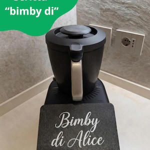 Op de afbeelding: Een zwart keukenapparaat met een zilveren handvat en een donkergrijze basis. Een donkergrijs rechthoekig bord voor het apparaat leest "Bimby di Alice" in witte letters. De tekst "Scritta 'bimby di'" staat in een groene vorm linksboven.