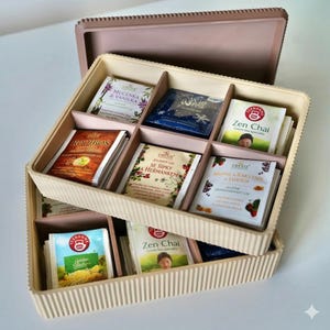 Puede incluir: Una caja de té beige con tapa, dividida en compartimentos llenos de bolsitas de té. Las bolsitas de té presentan envases coloridos con texto e imágenes, incluyendo "Rooibos" y "Zen Chai". La caja está apilada sobre otra caja similar.