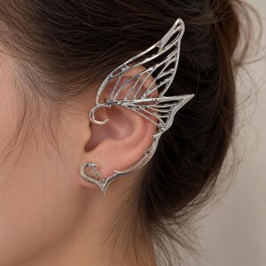 May include: Silver-toned ear cuff featuring a wing-like design. The cuff wraps around the ear with intricate, openwork detailing. The design includes a curved section that fits over the ear and a heart-shaped element at the bottom.