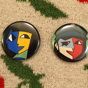 May include: Two round black buttons with abstract face designs. One features blue and yellow, the other red and blue. The buttons are on a textured, neutral-toned surface with green and red accents.