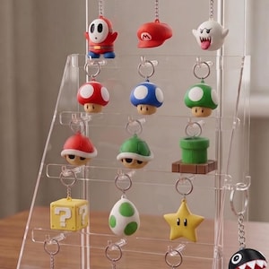 May include: A clear acrylic tiered display stand holding fourteen Super Mario themed keychains. The collection includes figures of Shy Guy, Mario's hat, Boo, various mushrooms, shells, a warp pipe, a question block, an egg, a star, and pixelated characters.