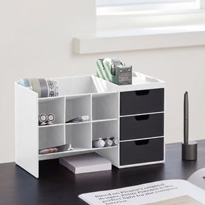 Op de afbeelding: Een witte bureau-organizer met een textuurafwerking, voorzien van zes open vierkante vakken, drie zwarte lades met uitgesneden handgrepen en een bovengedeelte met een groene tapehouder, diverse pennen, stiften en rollen tape.