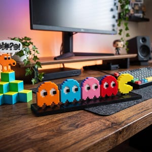 May include: A set of pixel art figures featuring an orange character on a green block pyramid with a speech bubble containing symbols, alongside a row of five colorful ghost characters and a yellow circular character on a black base.