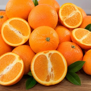 May include: A pile of fresh oranges, some whole and some halved, showcasing their vibrant orange color and juicy interior. Green leaves are scattered around the fruit, creating a natural and appealing arrangement. The oranges are on a wooden surface.