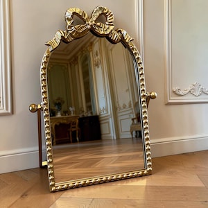 Op de afbeelding: Een goudkleurige, boogvormige tafelspiegel met een decoratieve strik aan de bovenkant en een frame met kralenrand. De spiegel rust op een standaard met twee knoppen aan de zijkanten, waardoor deze kantelbaar is. Het spiegeloppervlak toont een kamer.