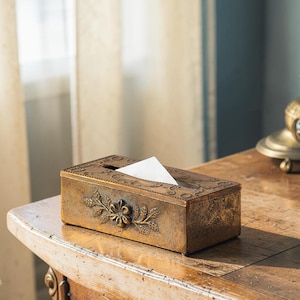 Könnte beinhalten: Ein rechteckiger Taschentuchbox-Halter mit einer patinierten Bronzeoberfläche. Die Außenseite zeigt ein geprägtes Blumenmotiv auf der Vorderseite und einen dekorativen Rand um die obere Öffnung, aus der ein weißes Taschentuch herausragt.